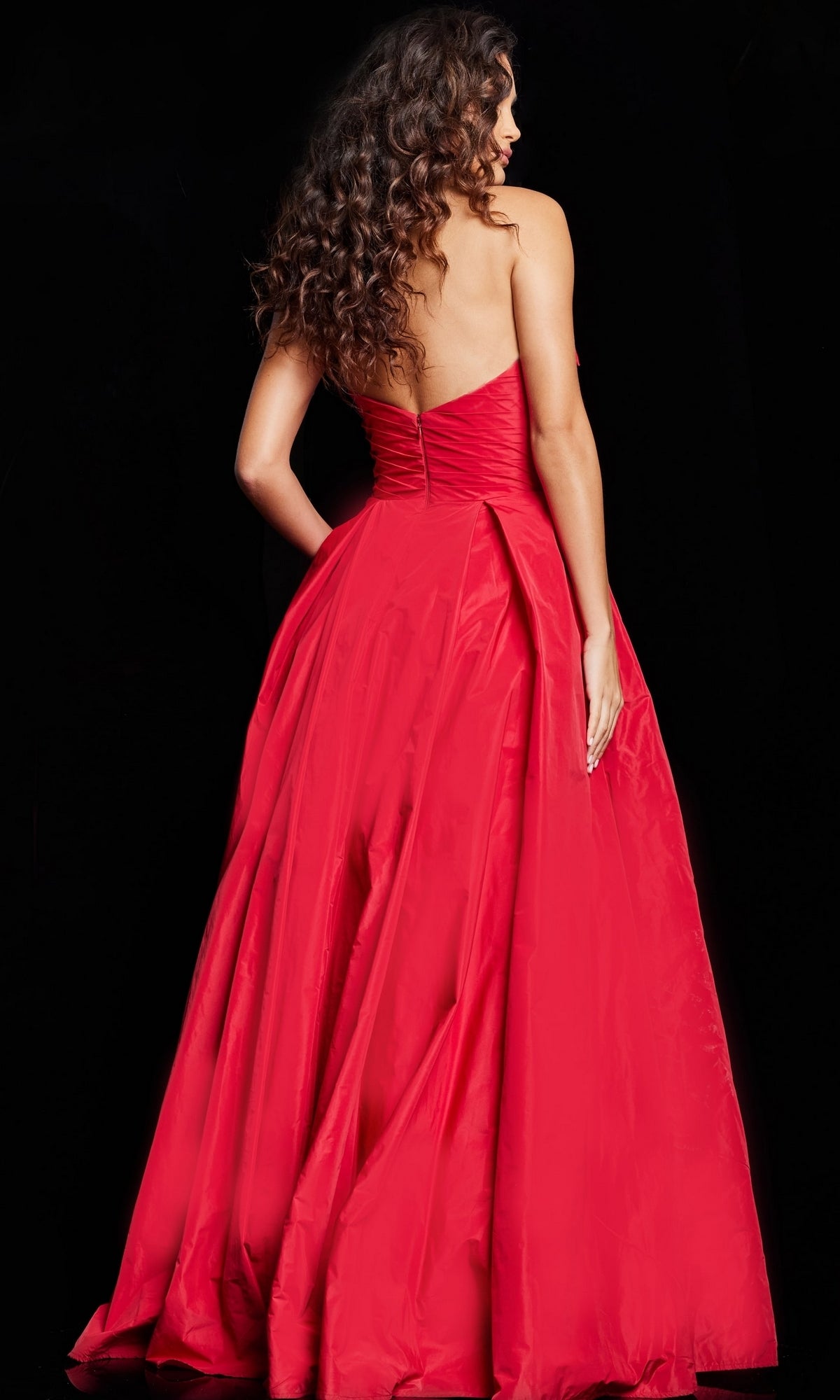 A woman with long curly hair is seen from the back, wearing the Long Prom Dress 37266 by Jovani—a floor-length, strapless red taffeta A-line gown—against a dramatic black background.